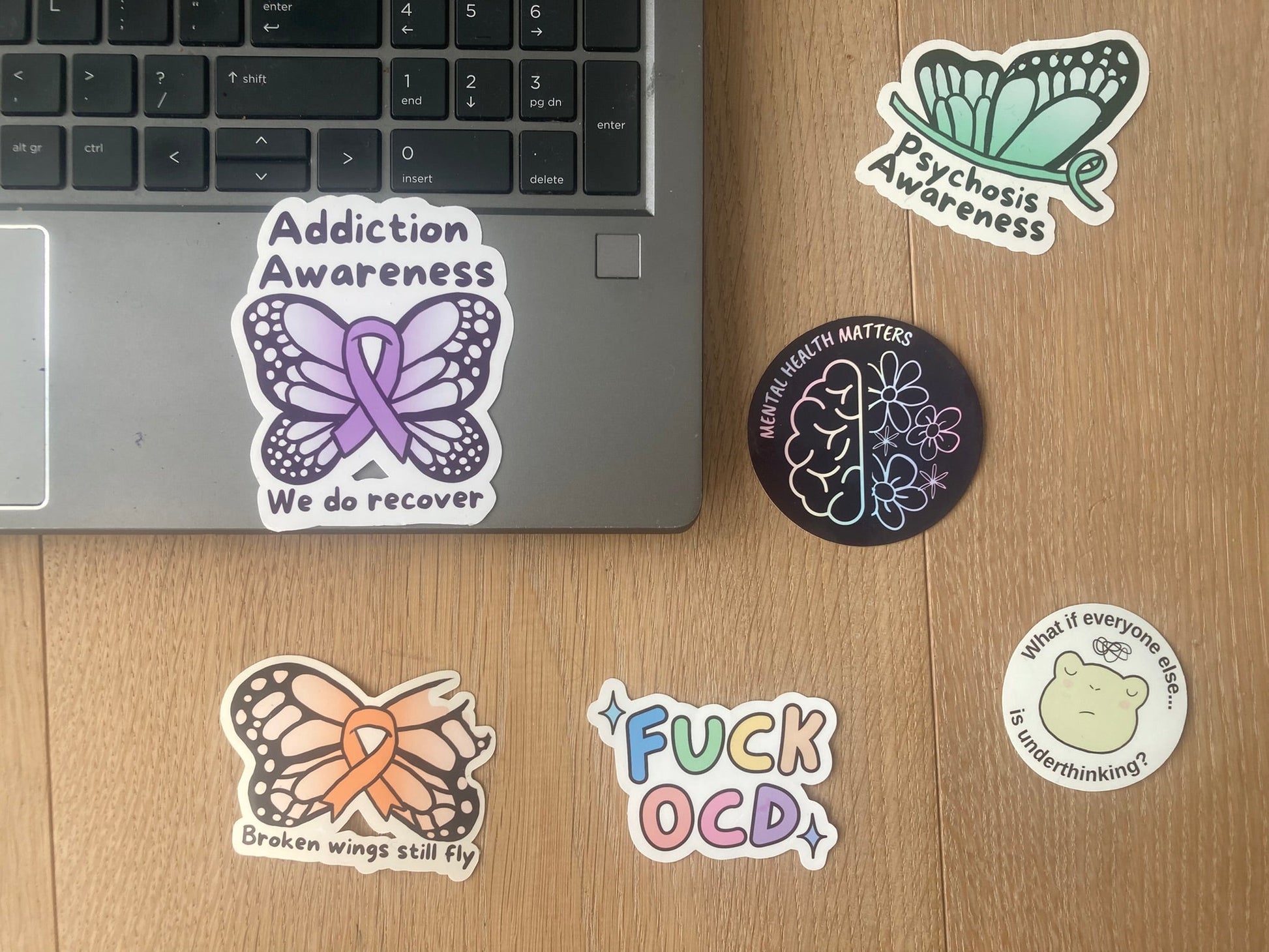 Addiction Awareness: we do recover shown as laptop sticker surrounded by other mental health stickers , handmade by NeurodiverseTea