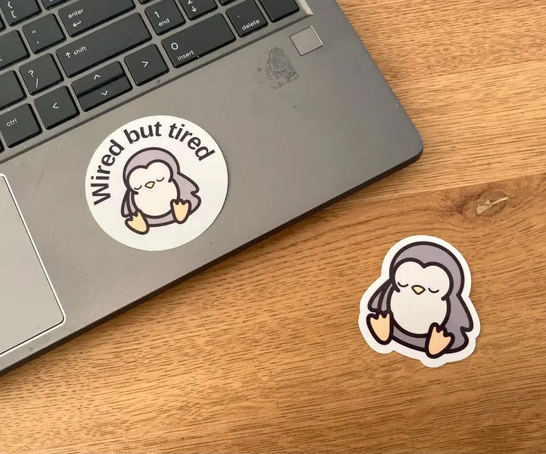 "Wired but tired" sleepy penguin stickers shown as laptop sticker, handmade by NeurodiverseTea